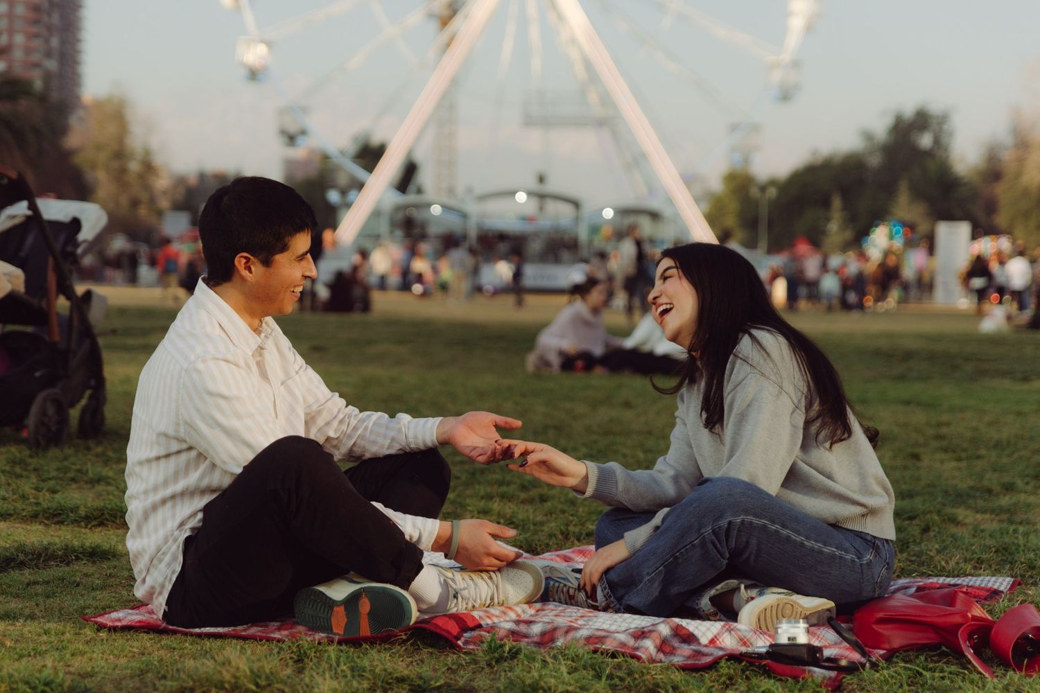 Smiling couple sharing snacks on a picnic blanket with a Ferris wheel and festival crowd behind them.
