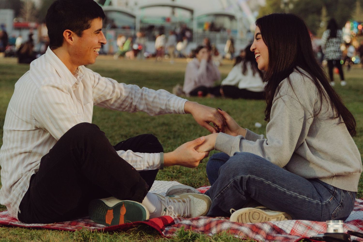 Laughing couple holding hands on a picnic blanket at an outdoor park festival during golden hour.