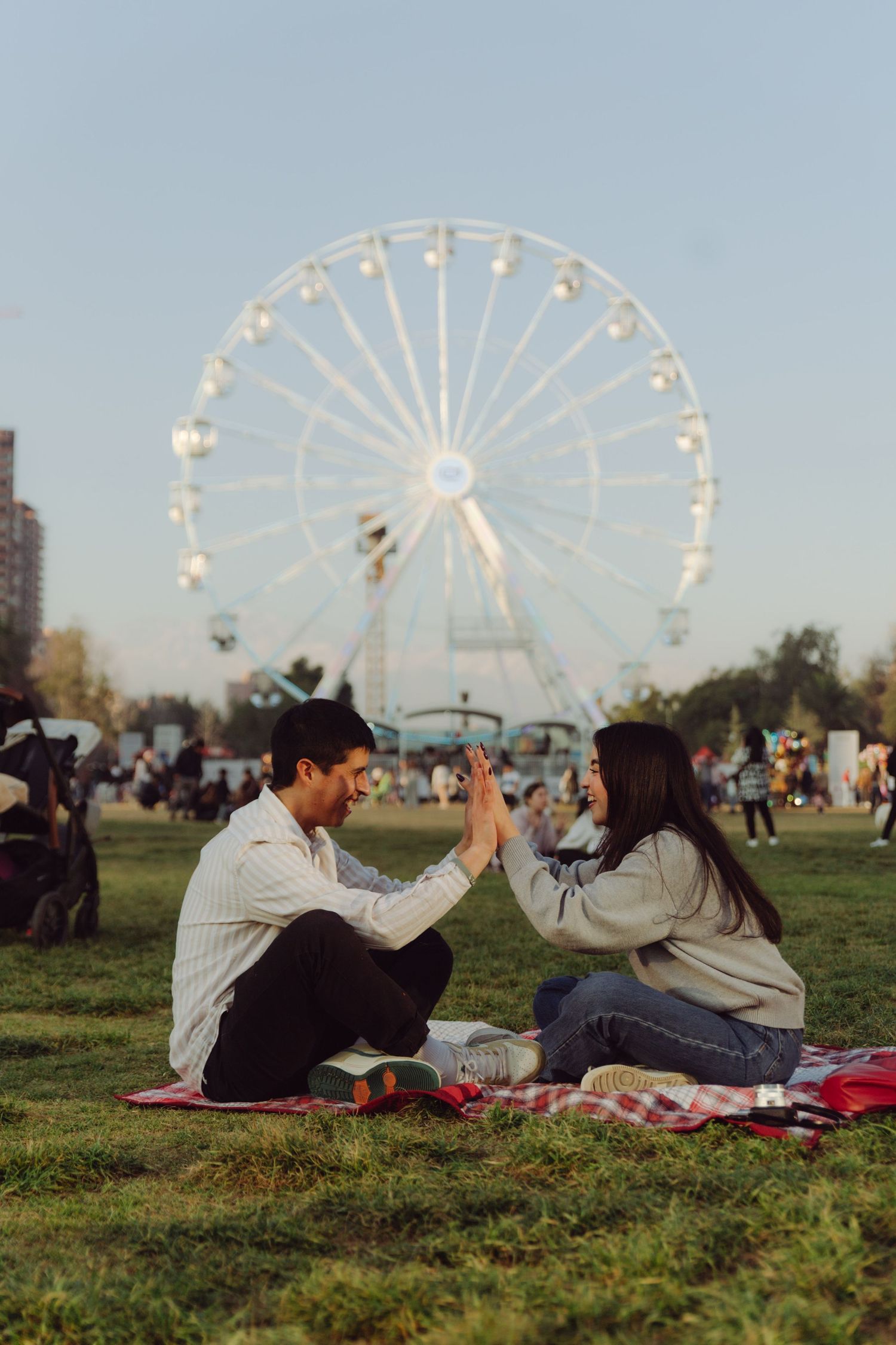 Joyful couple giving a high five on a picnic blanket with a Ferris wheel at sunset behind them.