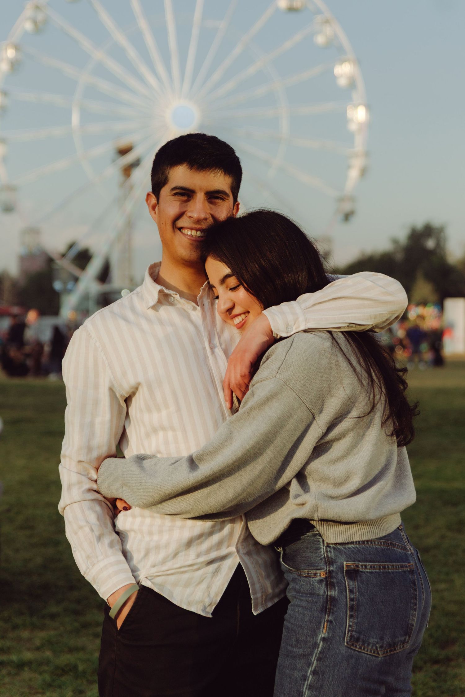 Smiling couple embracing in front of a ferris wheel at an outdoor fair during golden hour.