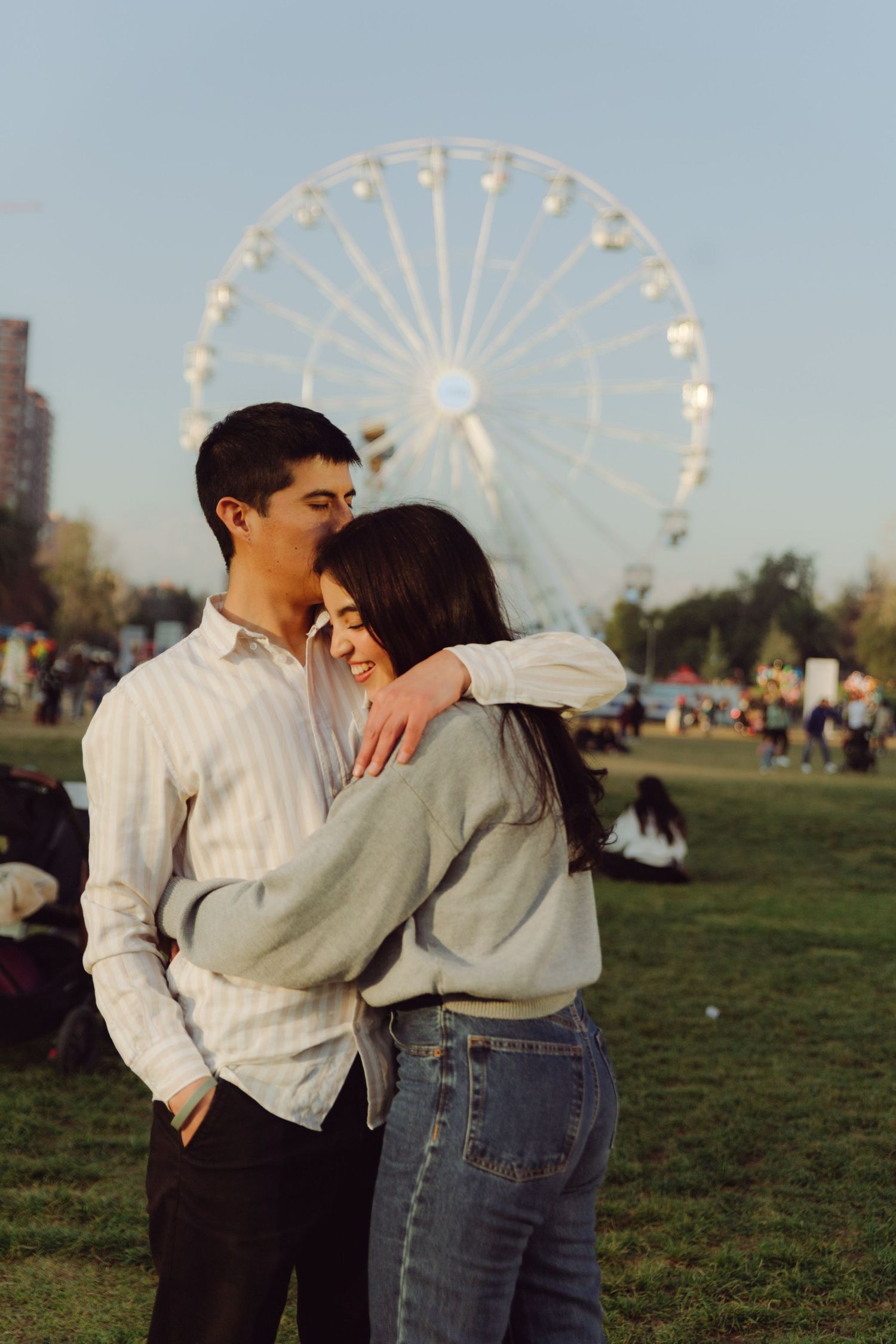 Romantic couple cuddling in a park with a large ferris wheel in the background.