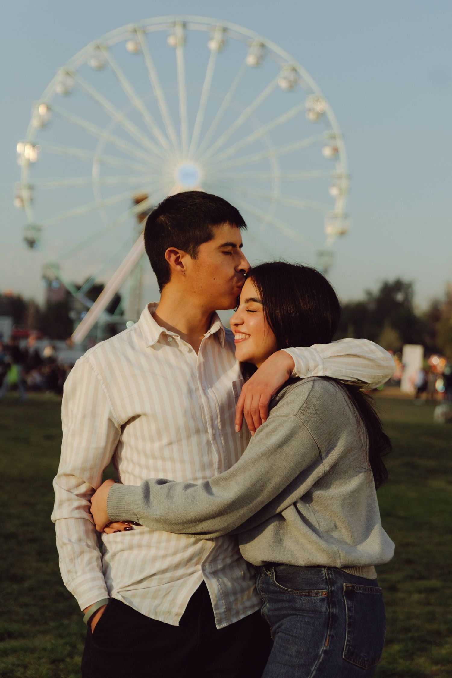 Young couple sharing a tender moment with a ferris wheel backdrop at an outdoor festival.