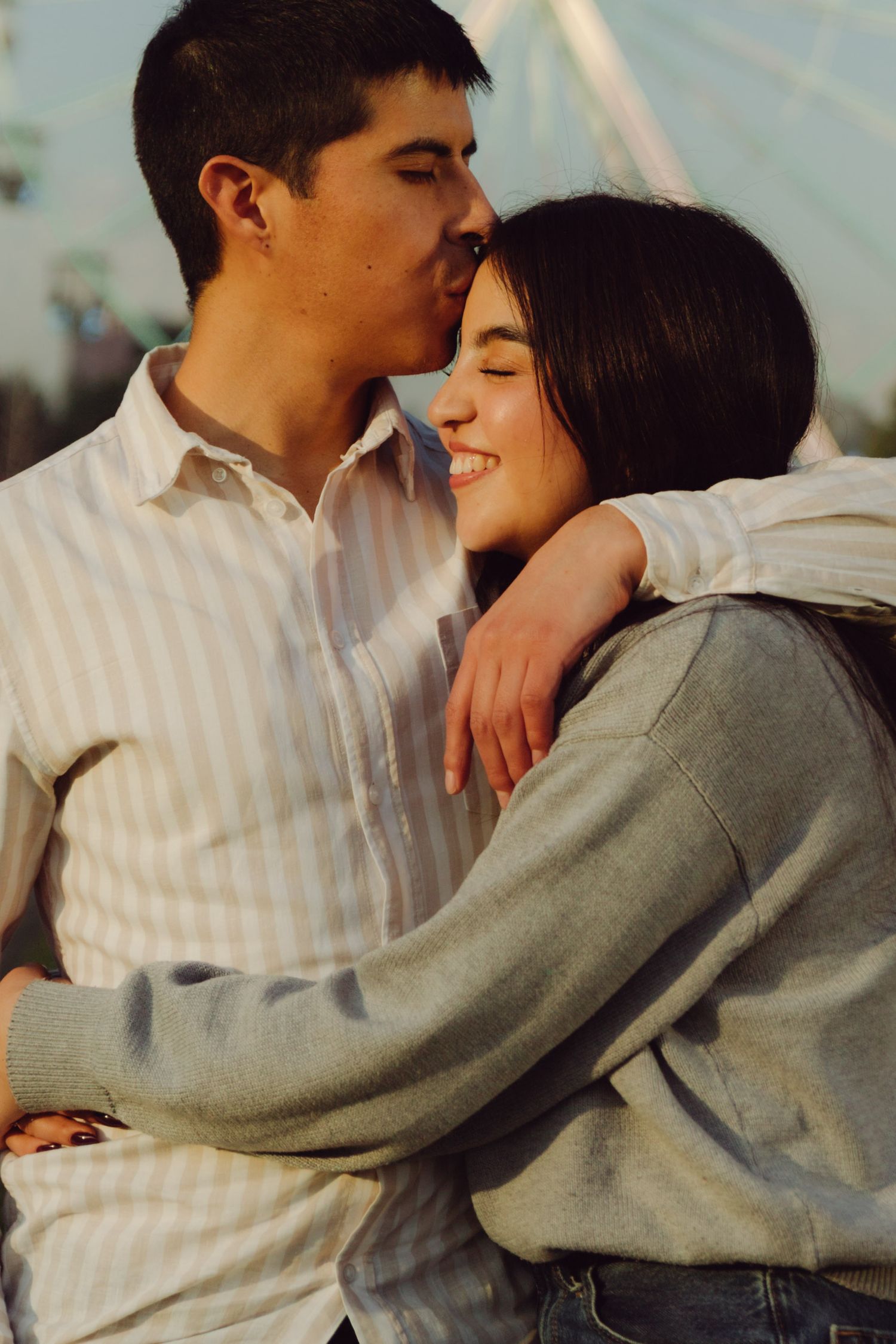 Man kissing woman on the cheek while embracing at an amusement park during golden hour.