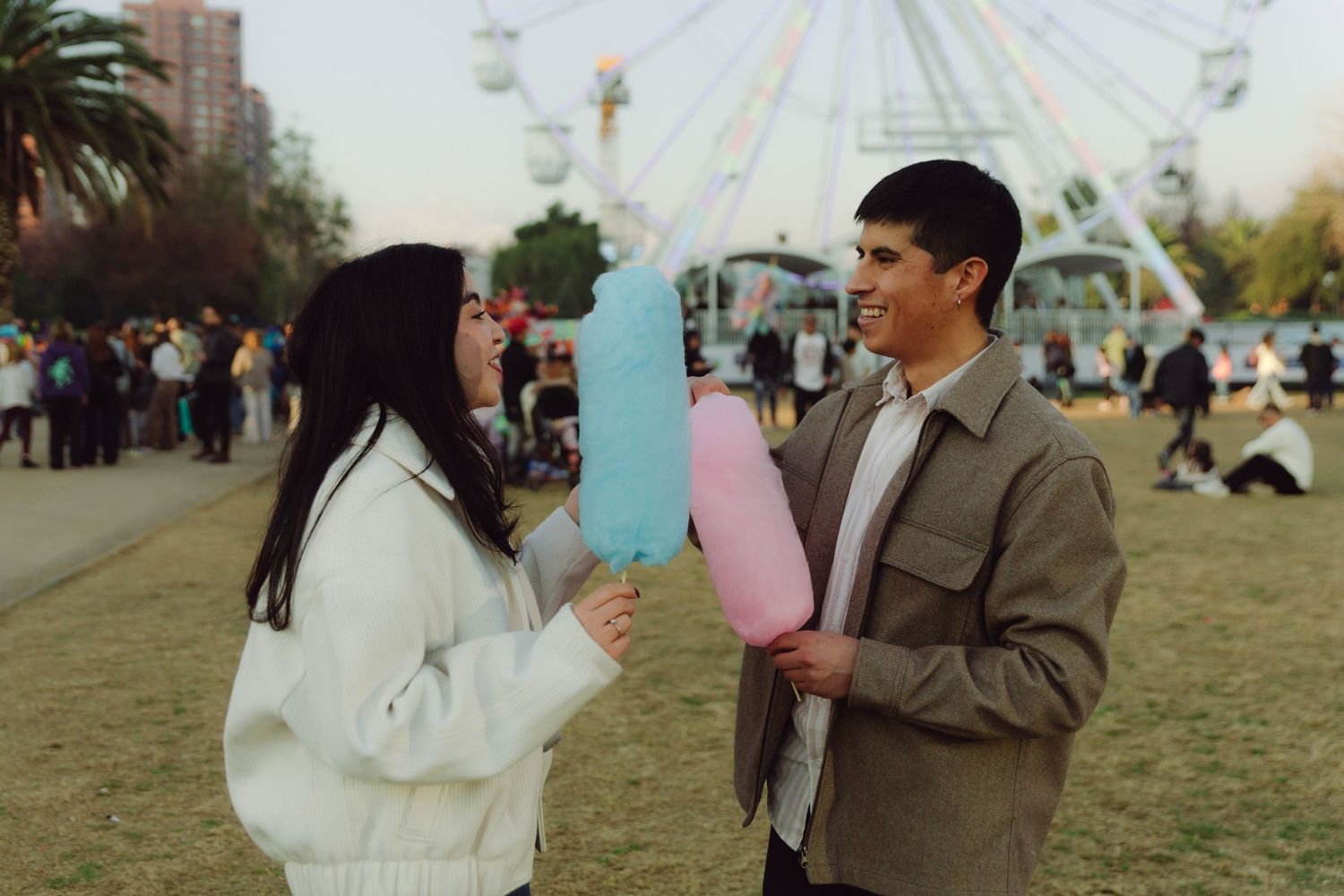Young couple holding blue and pink cotton candy at a festive outdoor fair near a ferris wheel.