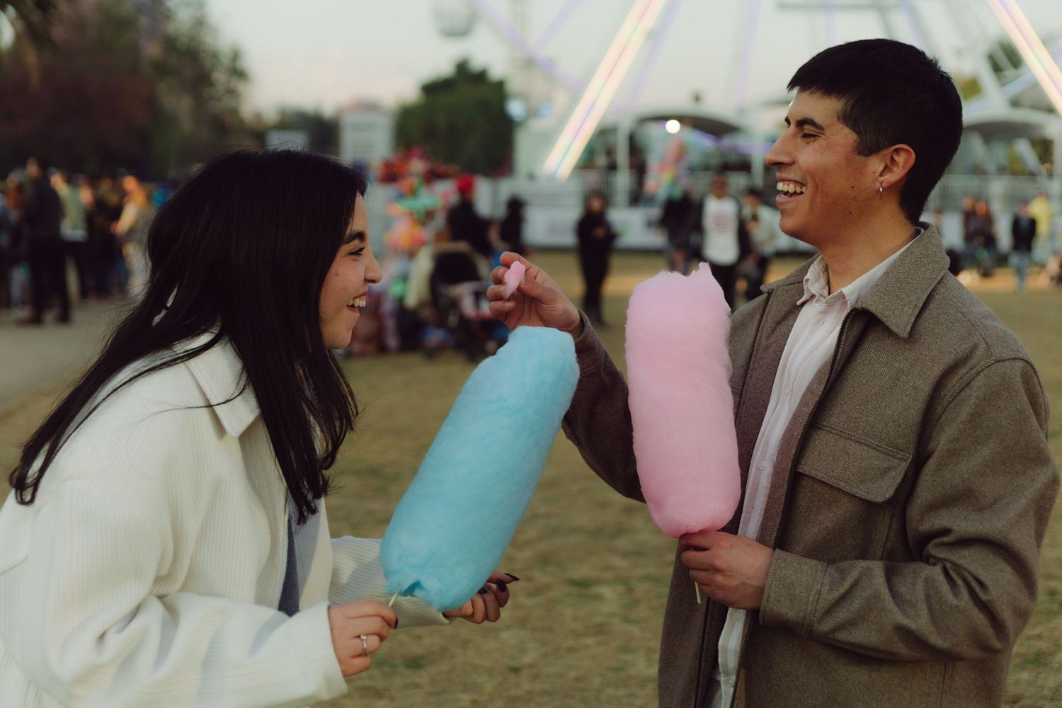 Smiling couple exchanging colorful cotton candy at an amusement park during dusk.