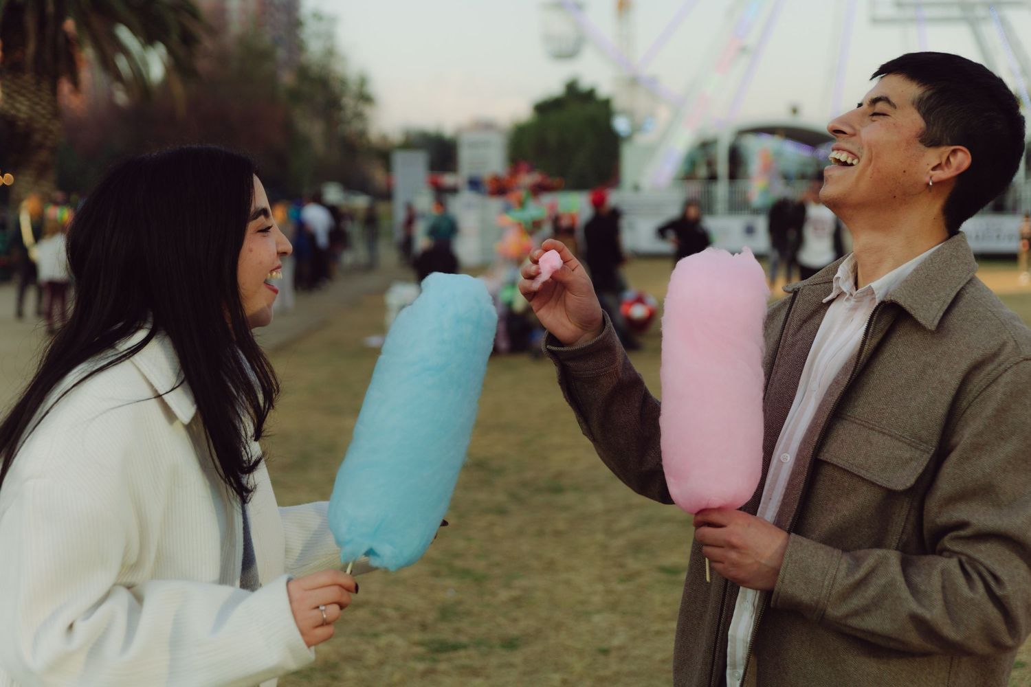 Happy couple laughing while holding blue and pink cotton candy at an outdoor fairground.