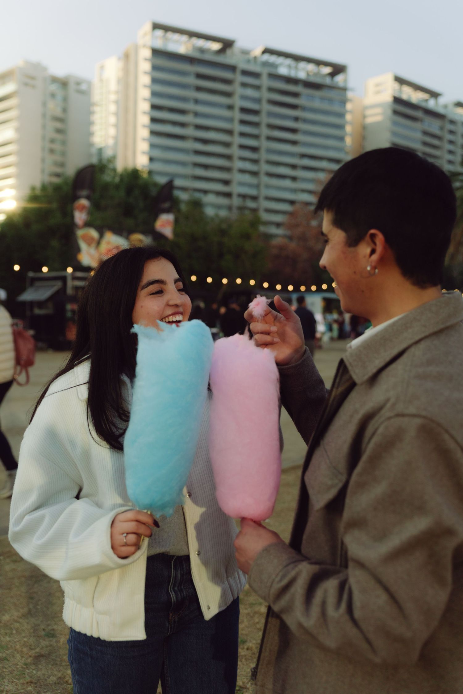 Joyful couple sharing cotton candy with city buildings and string lights in the background.