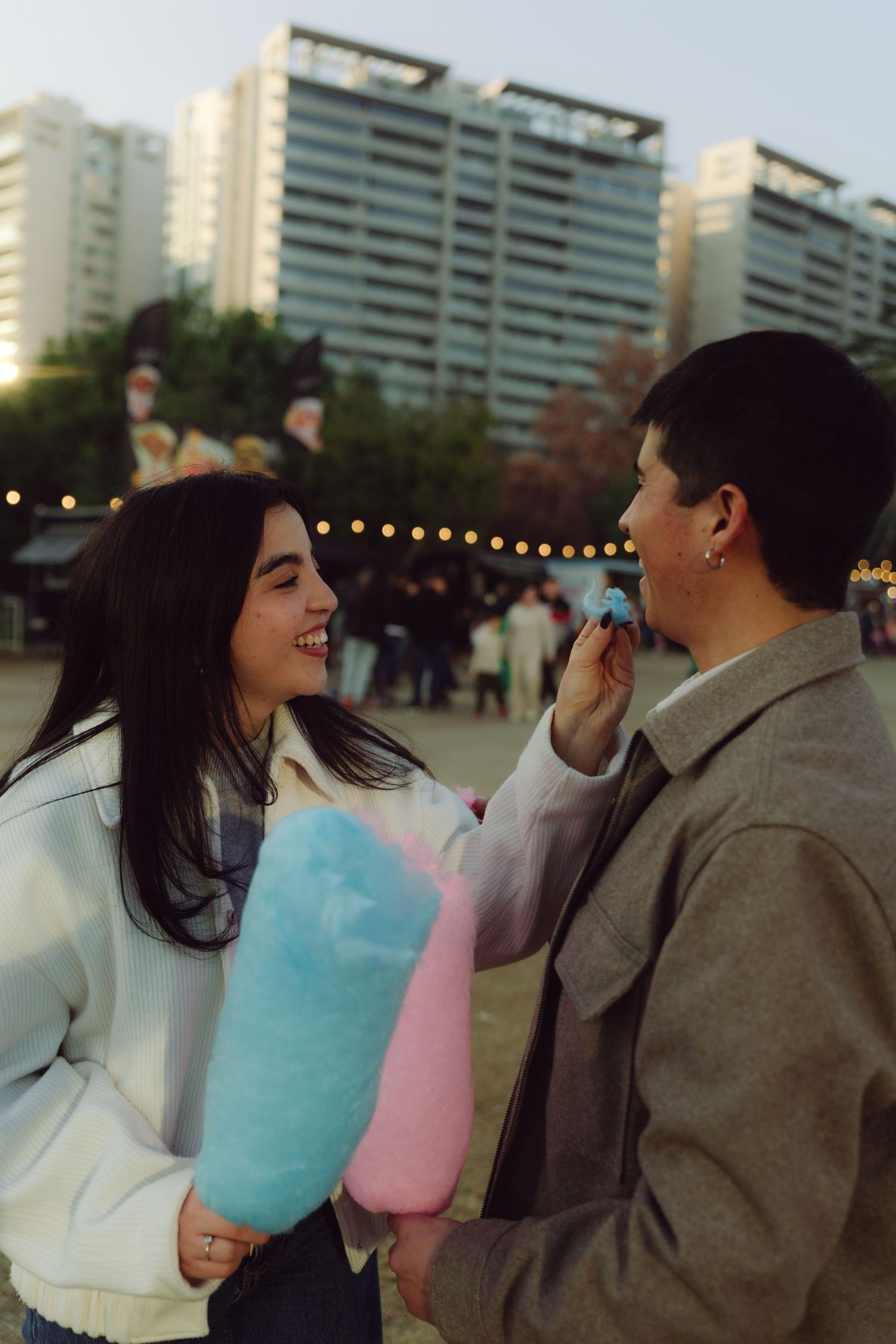 Young woman feeding her partner pink cotton candy at an outdoor evening festival.