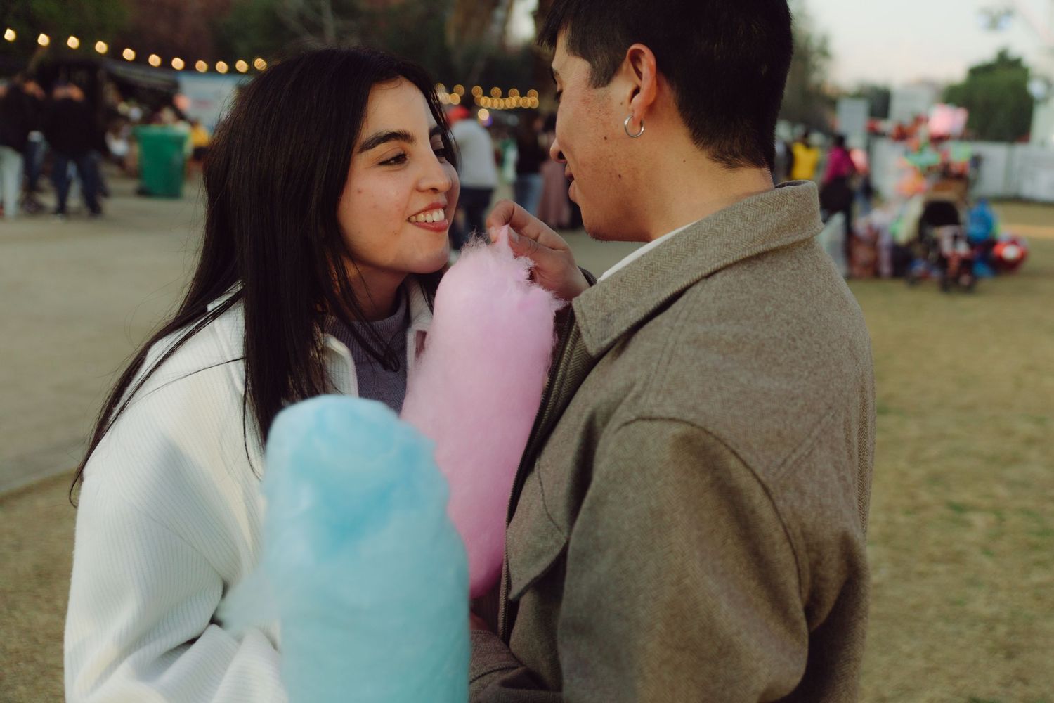Young couple sharing pink and blue cotton candy at a festive outdoor fair at dusk.