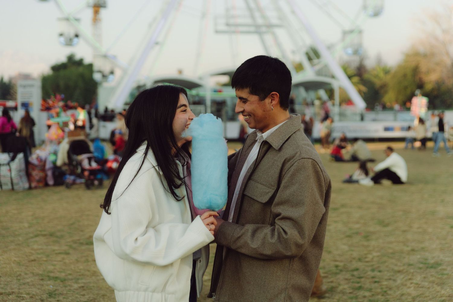 Smiling couple sharing blue cotton candy at an outdoor carnival with a Ferris wheel.