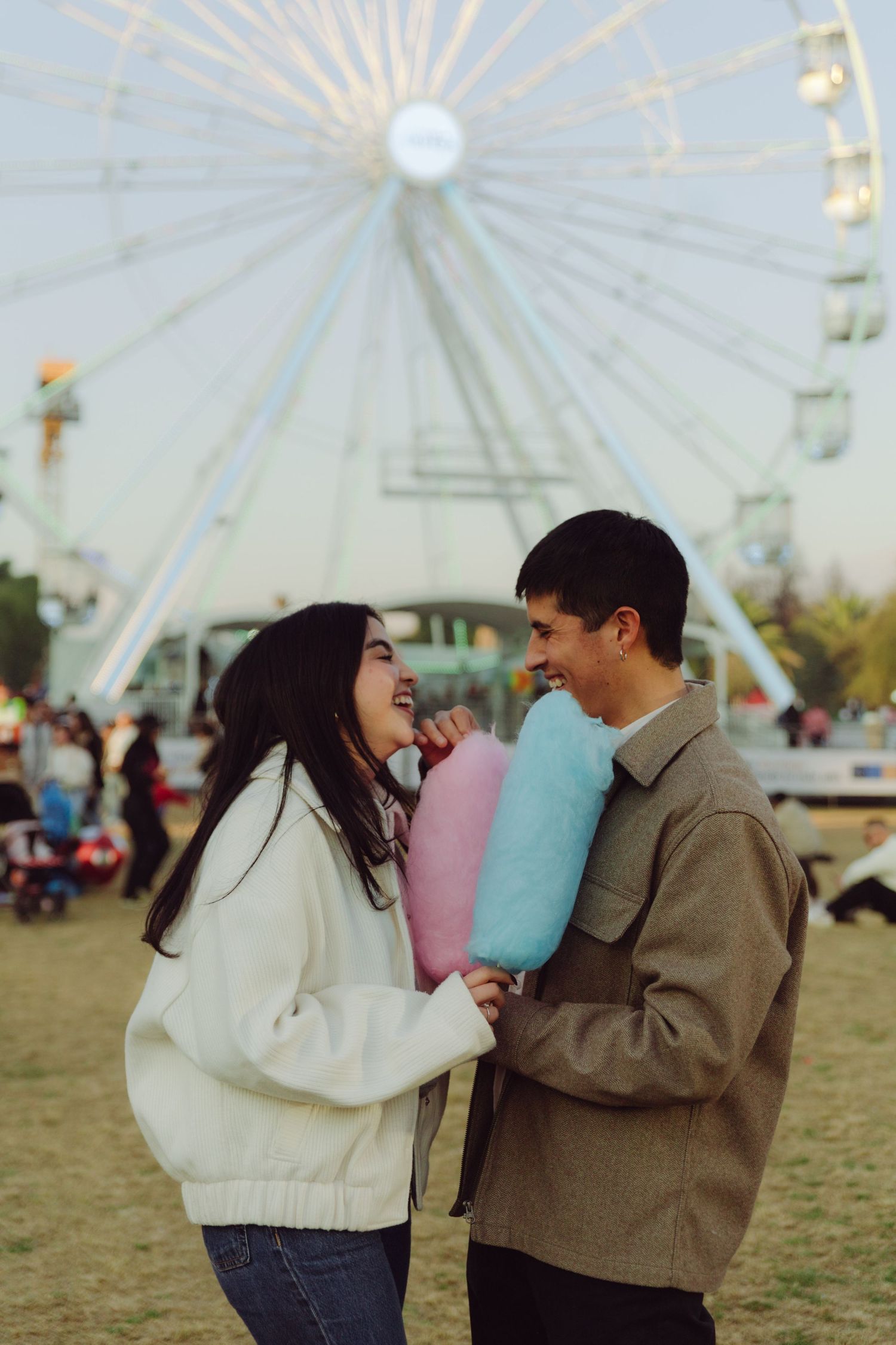 Joyful couple holding pink and blue cotton candy in front of a lit Ferris wheel.