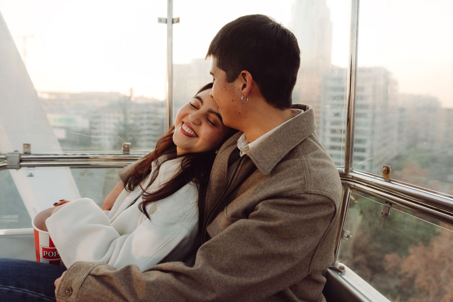 Happy couple cuddling inside a Ferris wheel gondola with city skyline views.