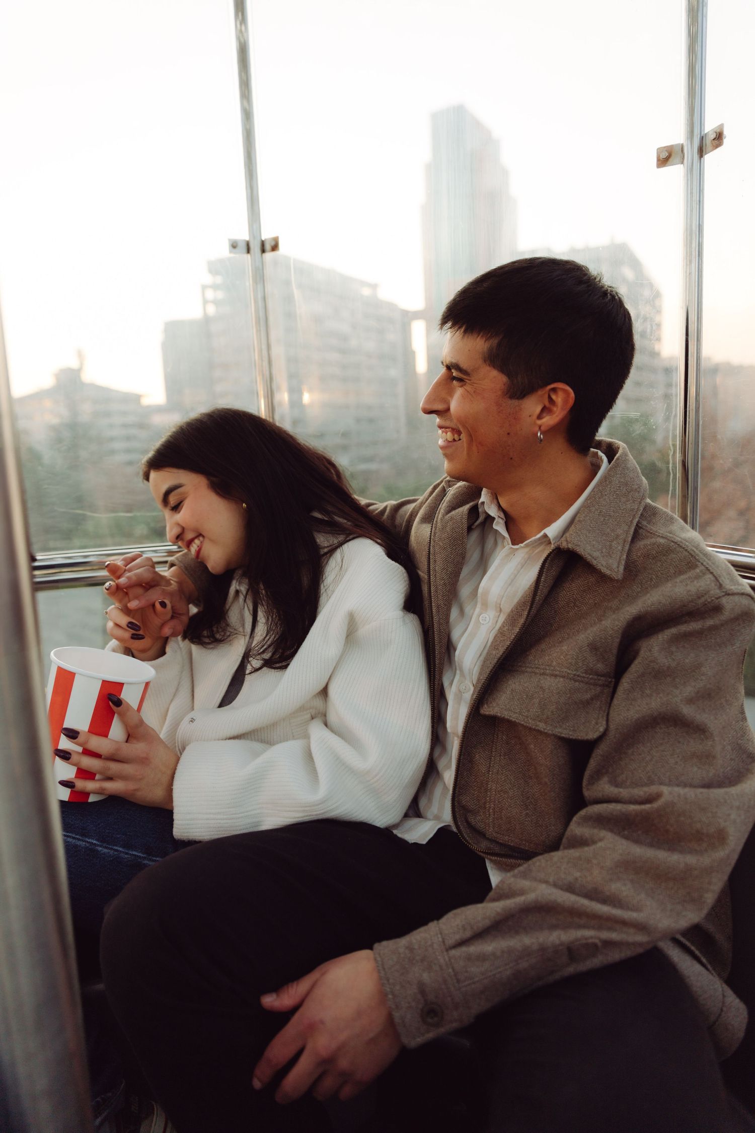 Smiling couple enjoying popcorn together inside a Ferris wheel overlooking a city.