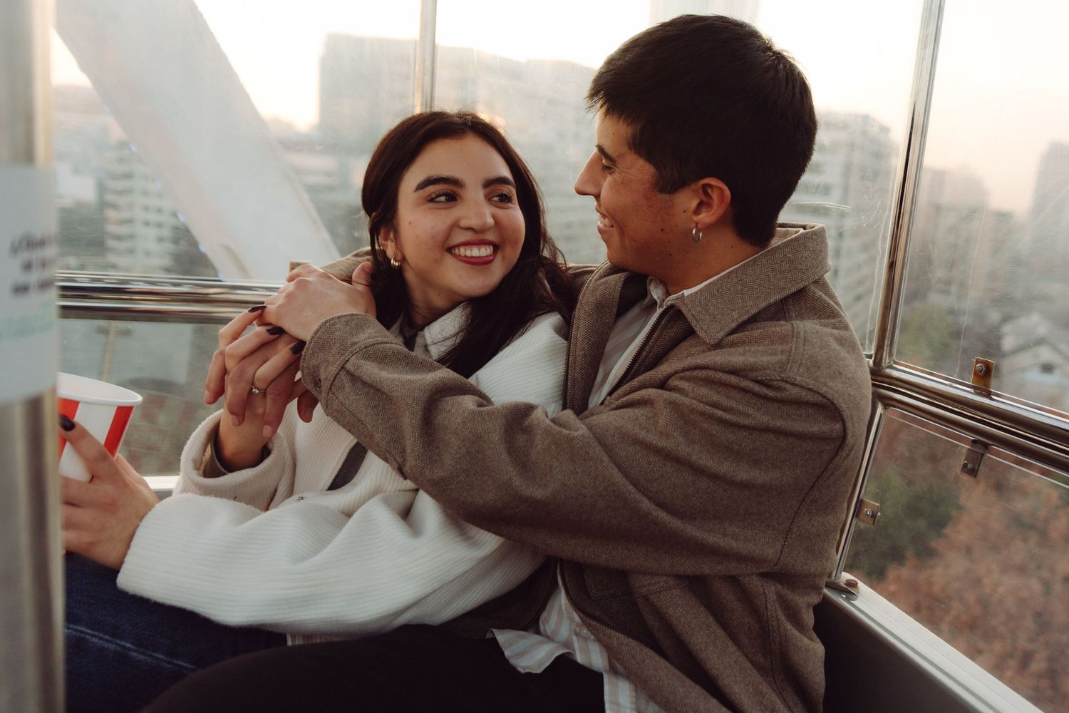 Romantic couple holding hands and smiling in a Ferris wheel gondola at sunset.