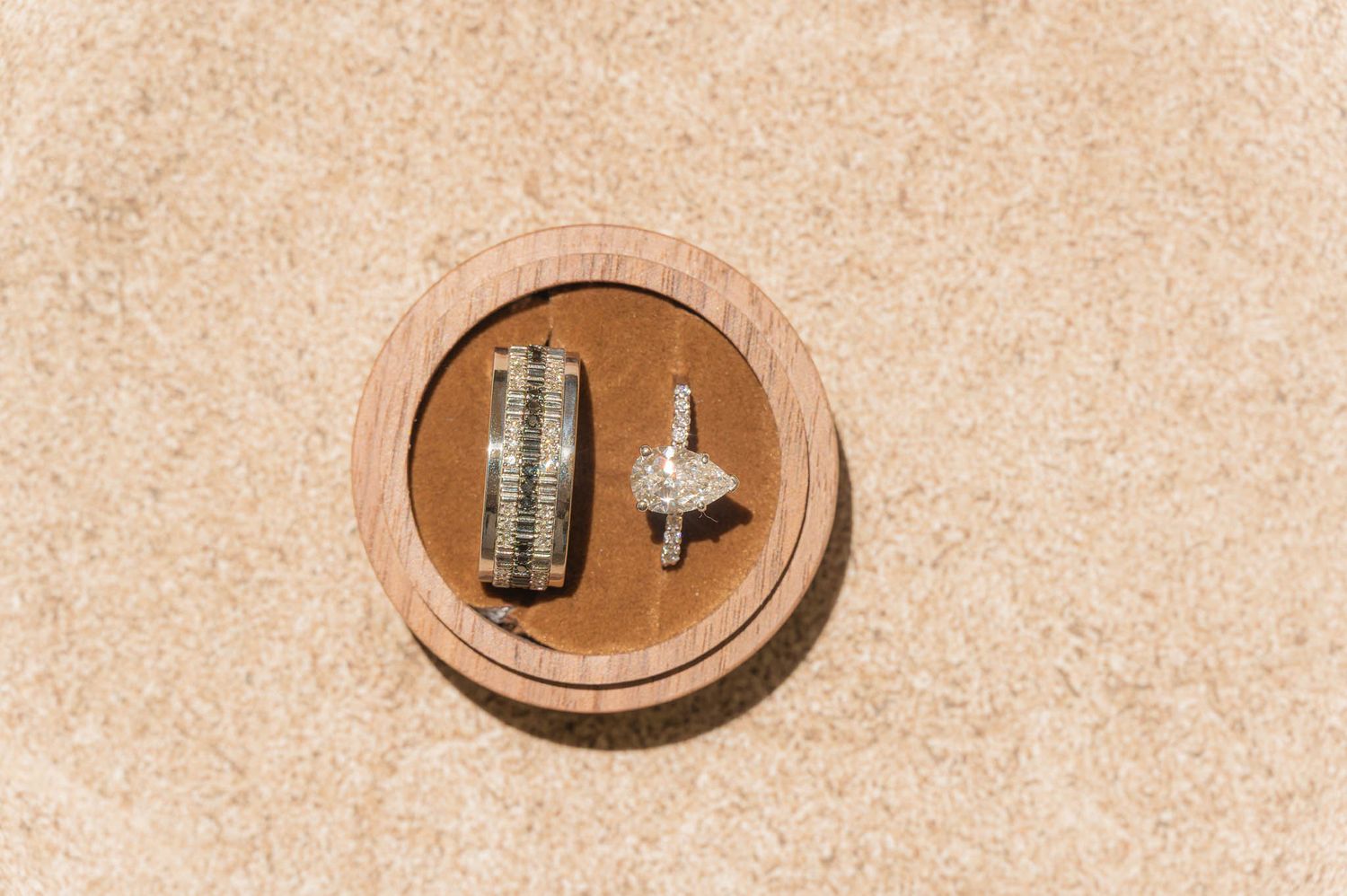 Two diamond wedding rings displayed in a round wooden ring box on a sandy beige surface.
