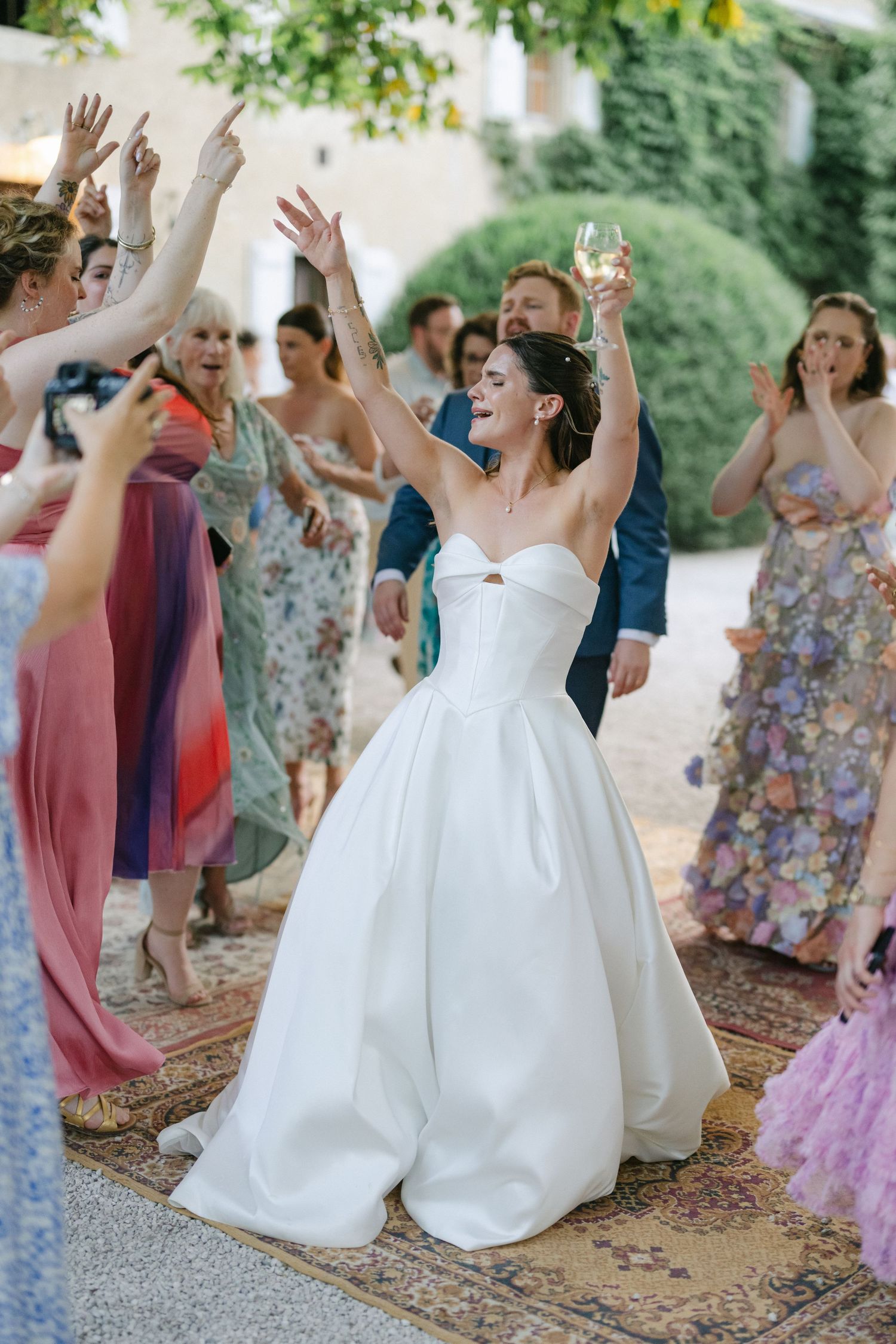 Bride and groom celebrate with cheering wedding guests outdoors, bride raising her arm joyfully in a white ballgown.