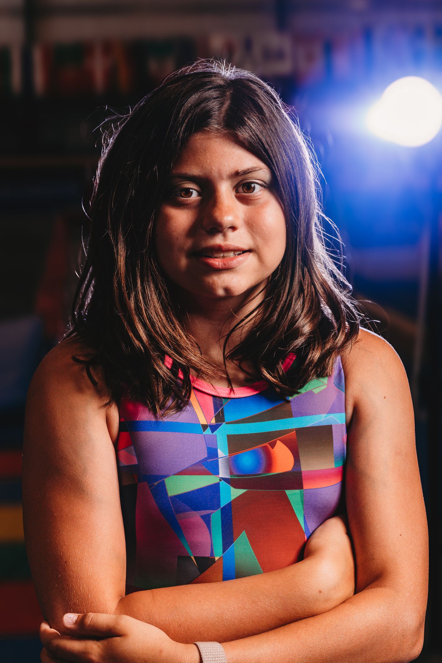 Young girl with dark hair in colorful geometric dress poses with arms crossed in front of stage lighting.
