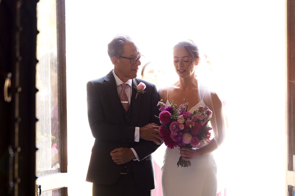 Bride in white gown holding pink bouquet walks with suited man through sunlit church doors.
