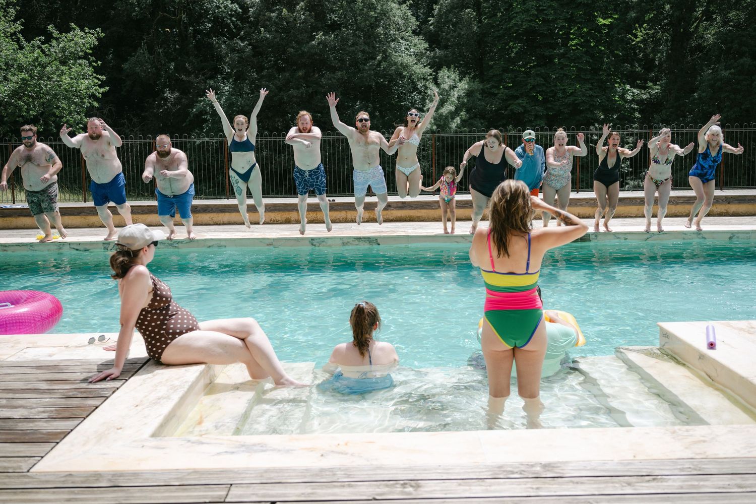 Group of friends jumping into outdoor pool on sunny day, with others watching and cheering from poolside deck.