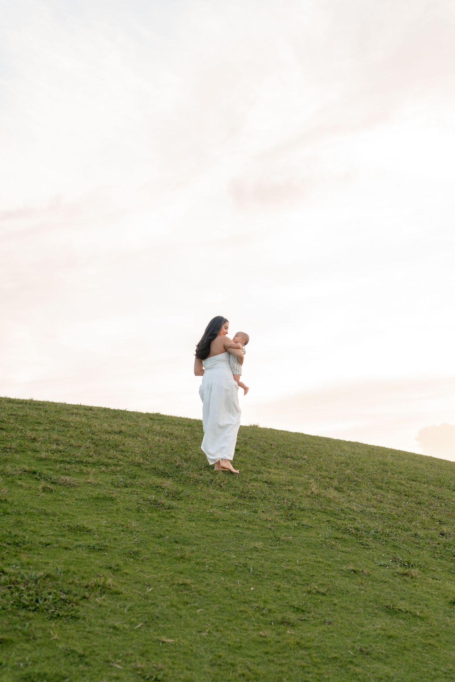 A woman in a white dress holds a baby on a lush green hillside under a soft cloudy sky.