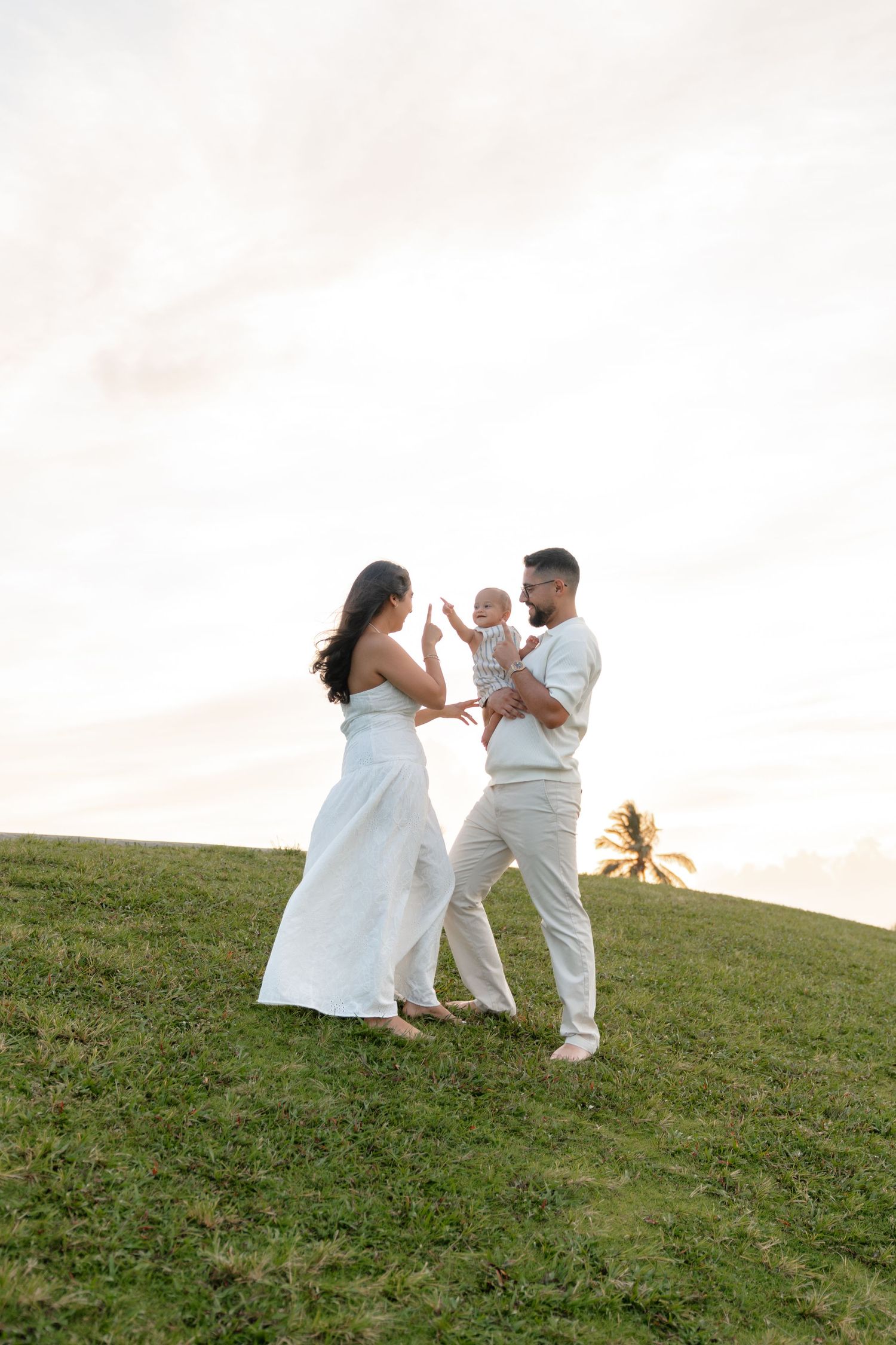 A happy family of three stands on a grassy hill at sunset, parents in white outfits holding their baby.