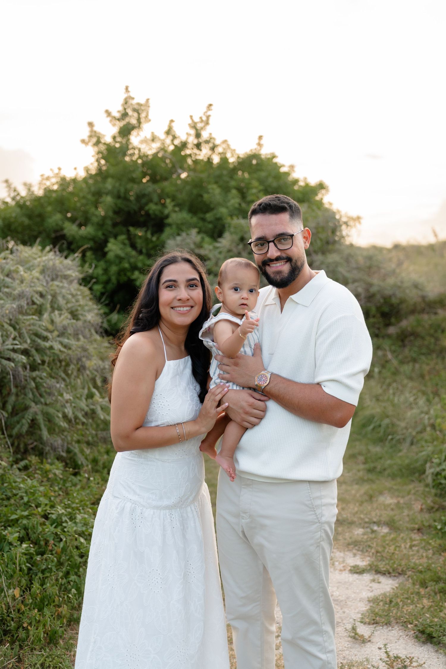 A smiling family of three poses outdoors, parents in white outfits holding their baby amid lush greenery at sunset.