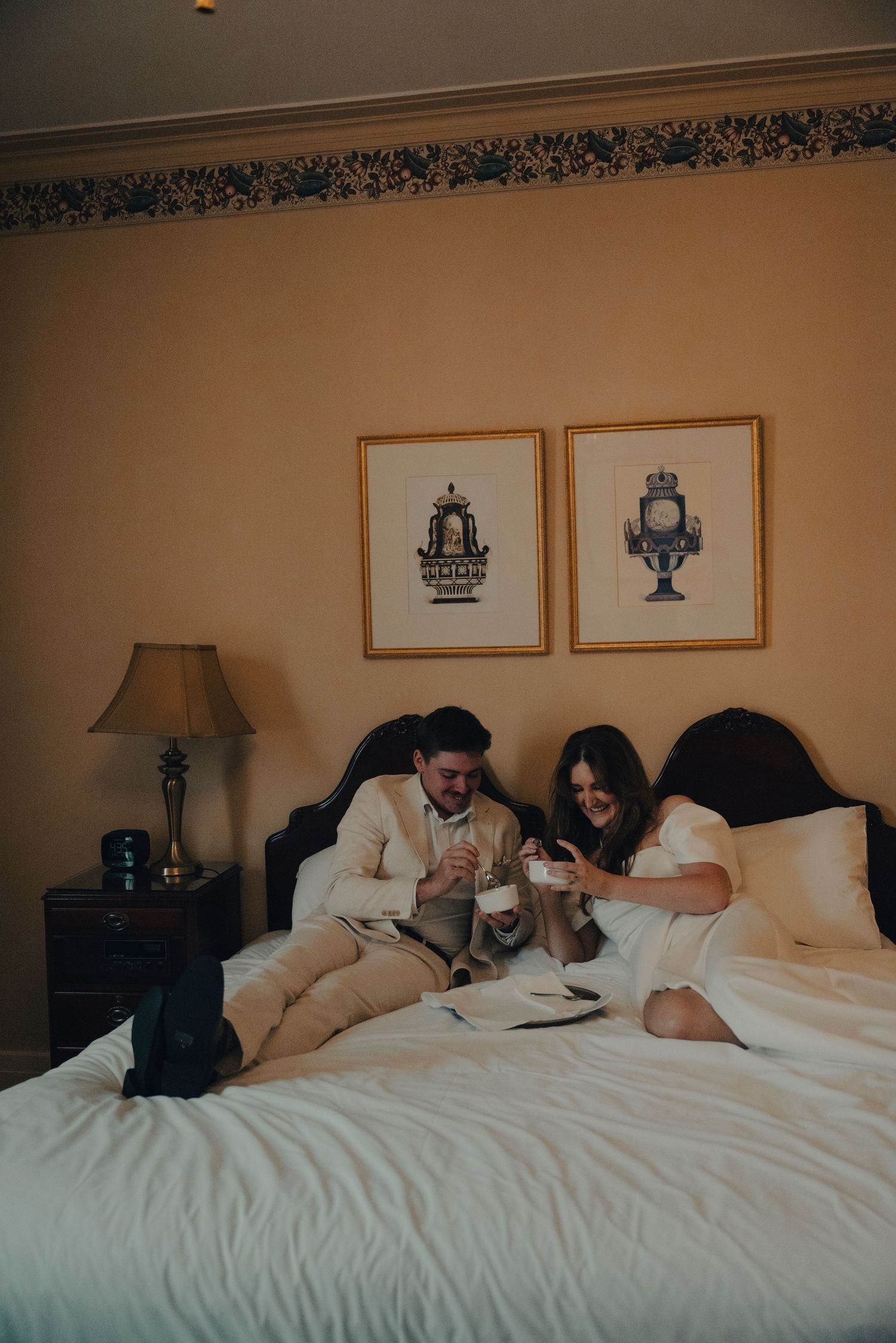 Couple relaxing on a hotel bed in elegant attire, enjoying drinks with framed artwork on beige walls.