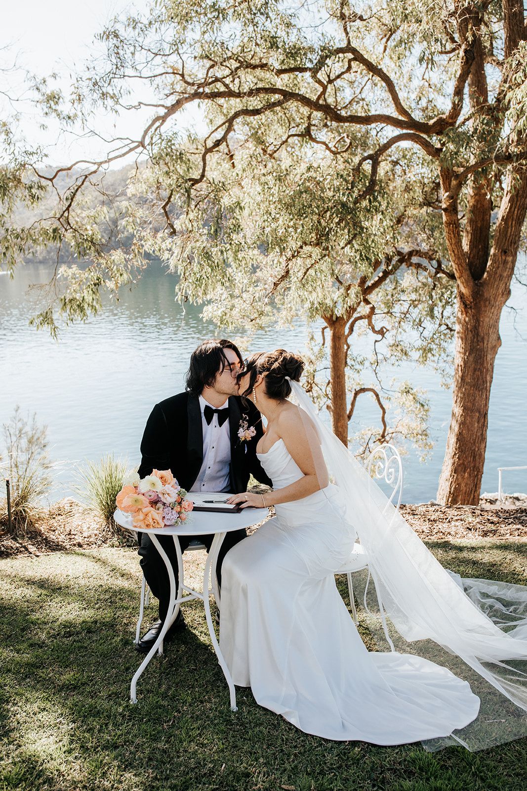 Bride and groom kissing at white table by serene lake under tree, bride in white gown with veil, groom in black tuxedo.
