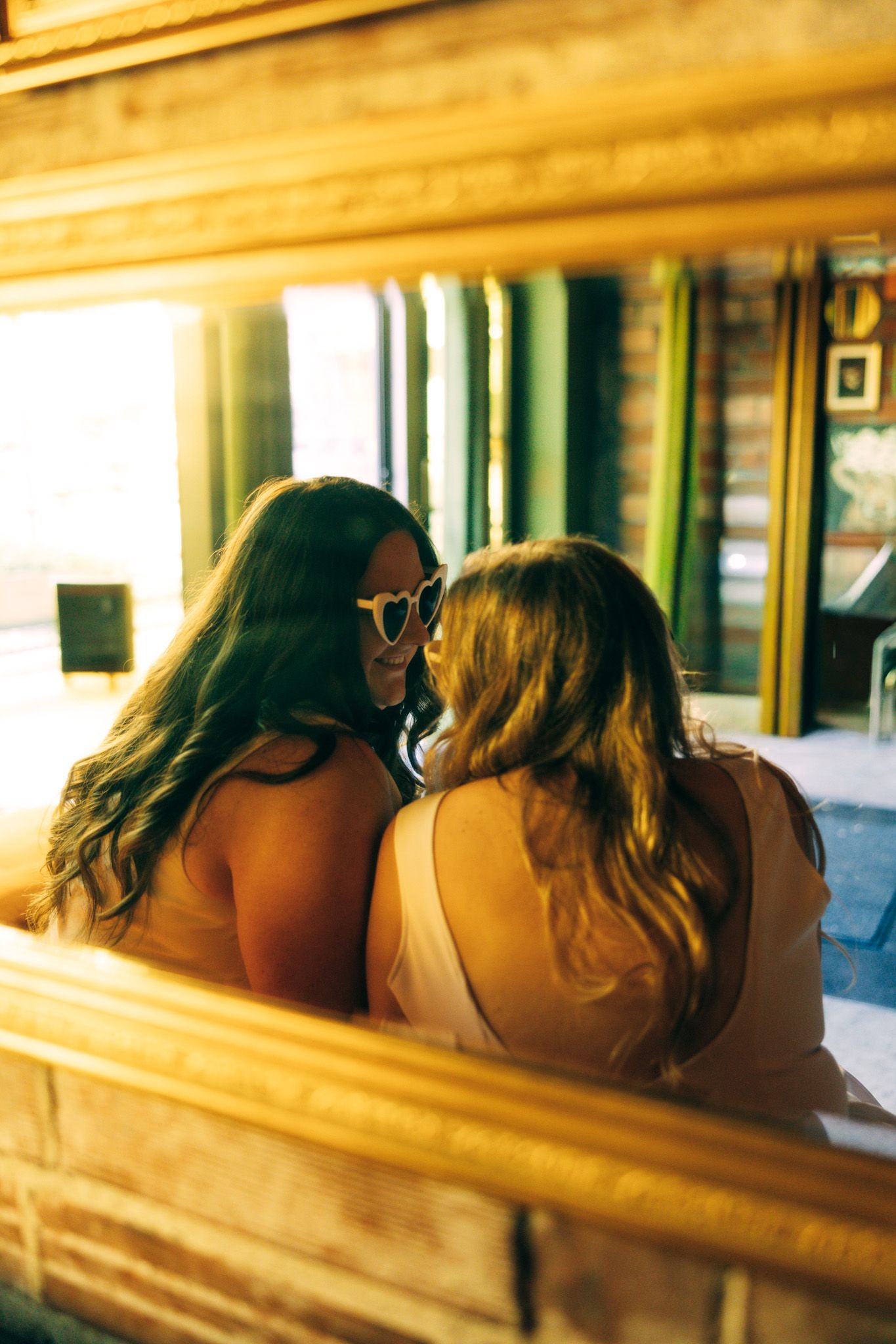 Two women with long hair sit closely together in warm golden light, viewed from behind in a rustic interior.