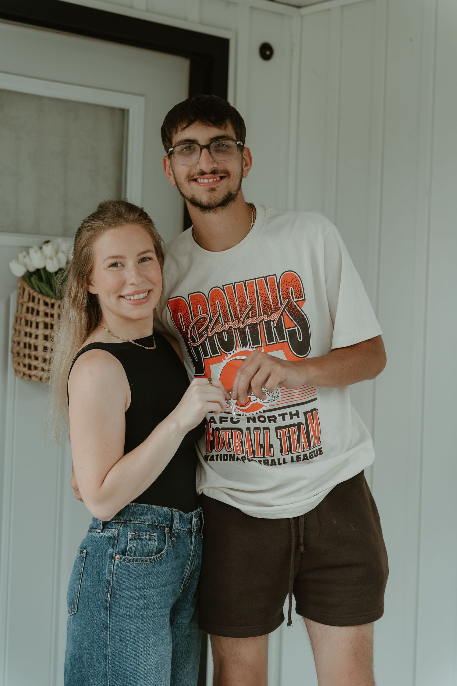 A smiling young couple posing together on a porch, man wearing a Cleveland Browns NFL t-shirt and dark shorts.