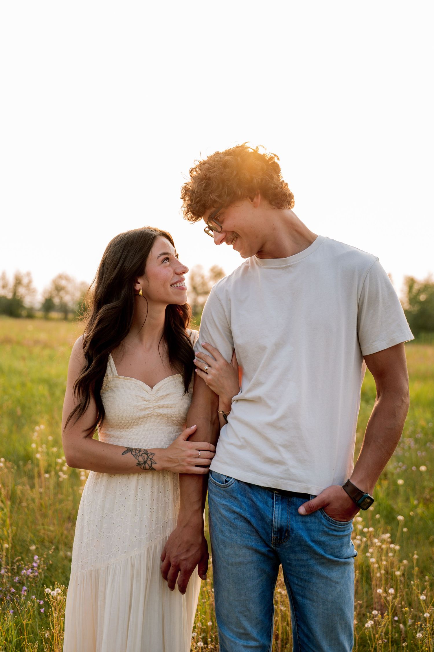 A smiling couple stands in a sunny meadow, the woman in a white dress and man in a white tee and jeans.