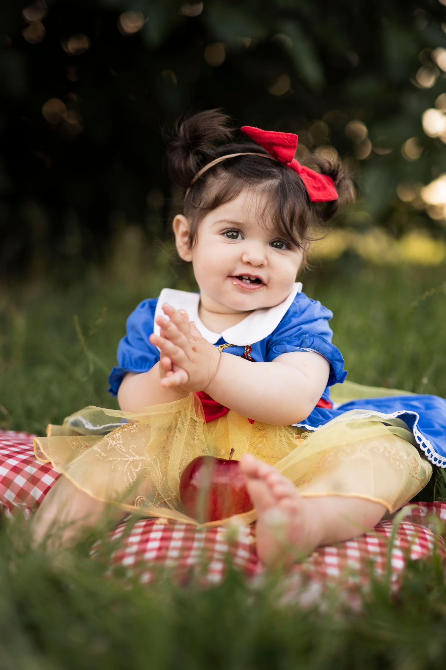 Baby girl in Snow White costume with red bow sits on checkered blanket outdoors with a red apple.