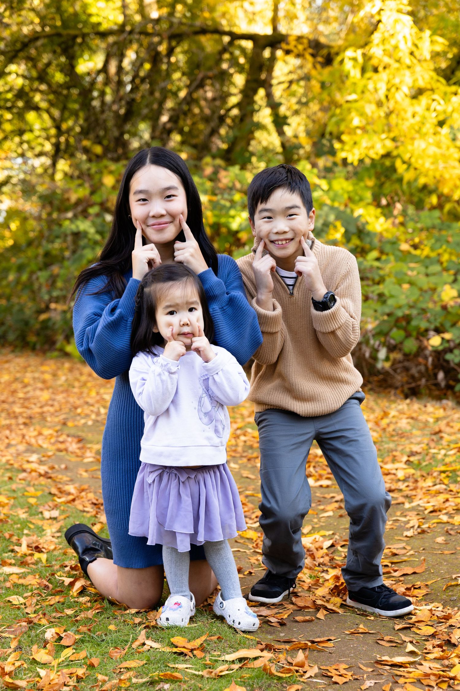 Three Asian children pose playfully in an autumn park, pointing at their smiles among golden fallen leaves.