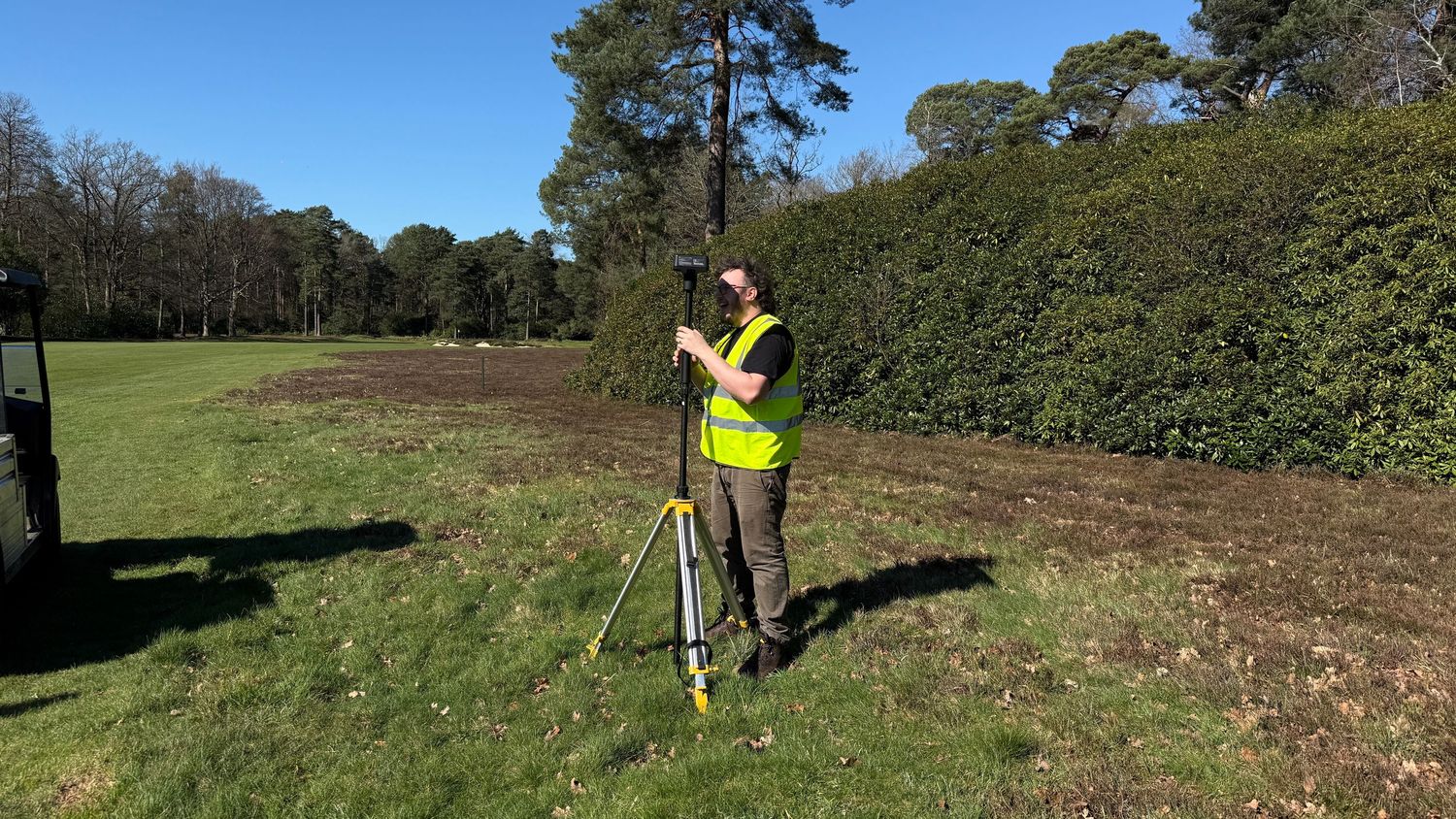 Surveyor in high-visibility vest using a tripod instrument on a green field surrounded by trees and hedges.