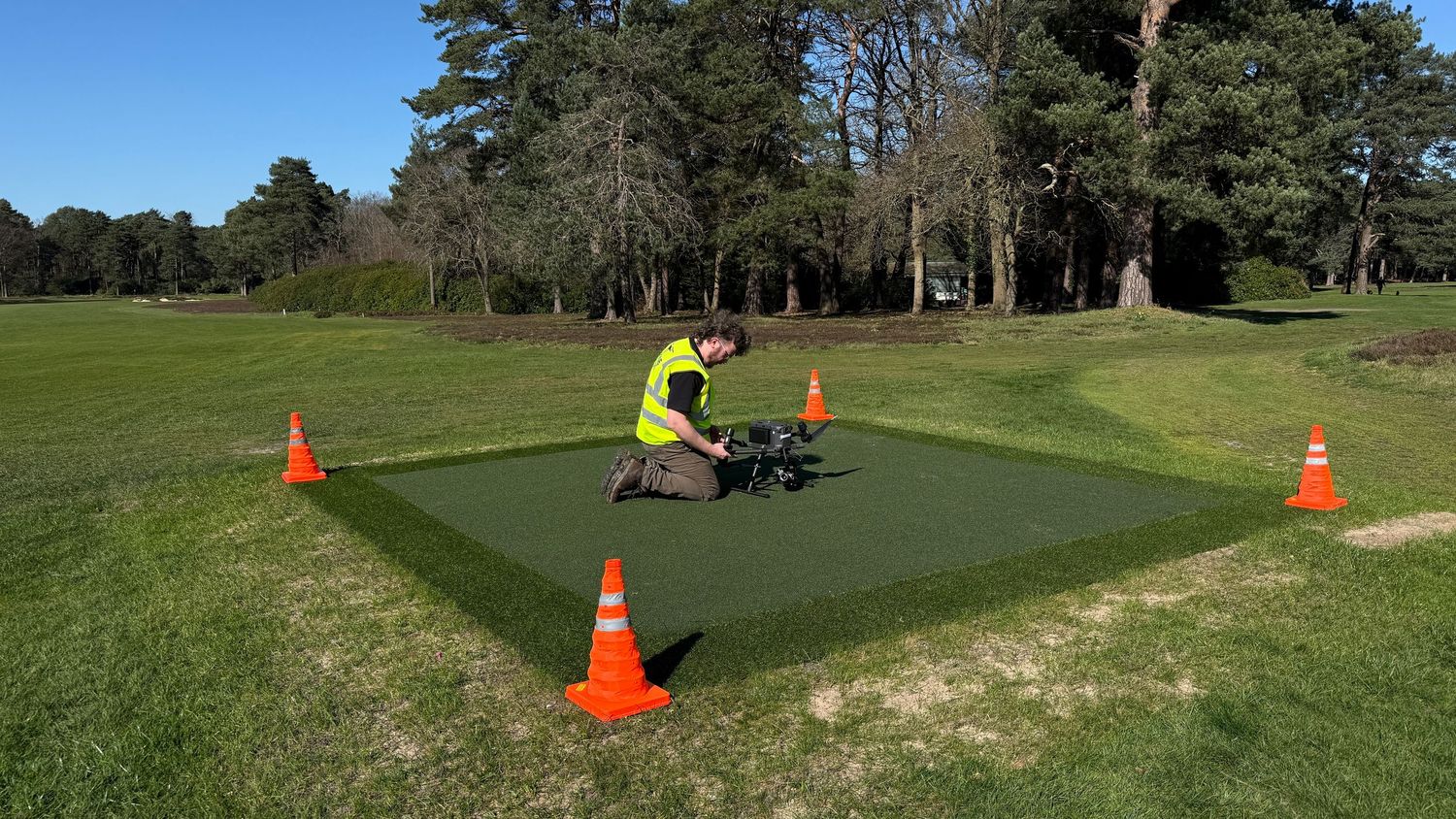 Drone operator in hi-vis vest kneeling on artificial turf landing pad marked with orange safety cones in a park.