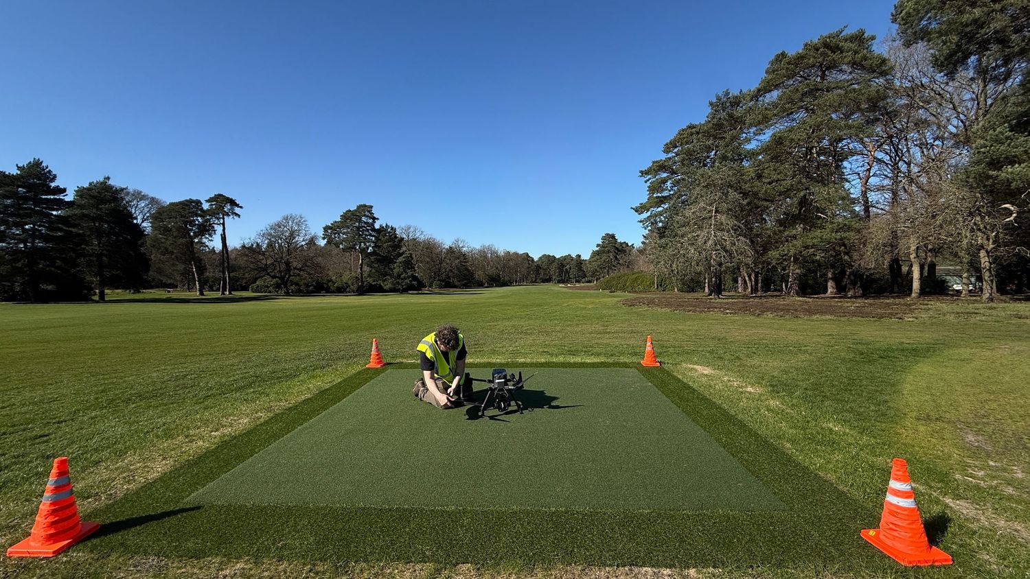 Golf course worker maintaining artificial turf driving range mat surrounded by orange cones on sunny day.