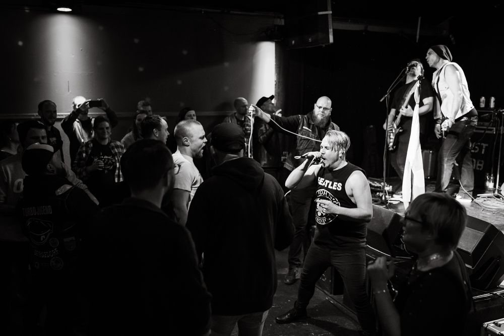 Black and white photo of a punk rock band performing live on stage with an energetic crowd in a dark venue.
