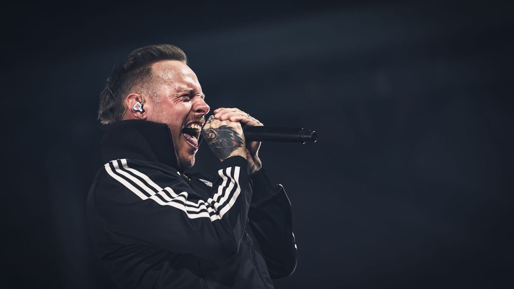 Tattooed male vocalist performing live on stage, singing passionately into microphone wearing black Adidas tracksuit jacket.