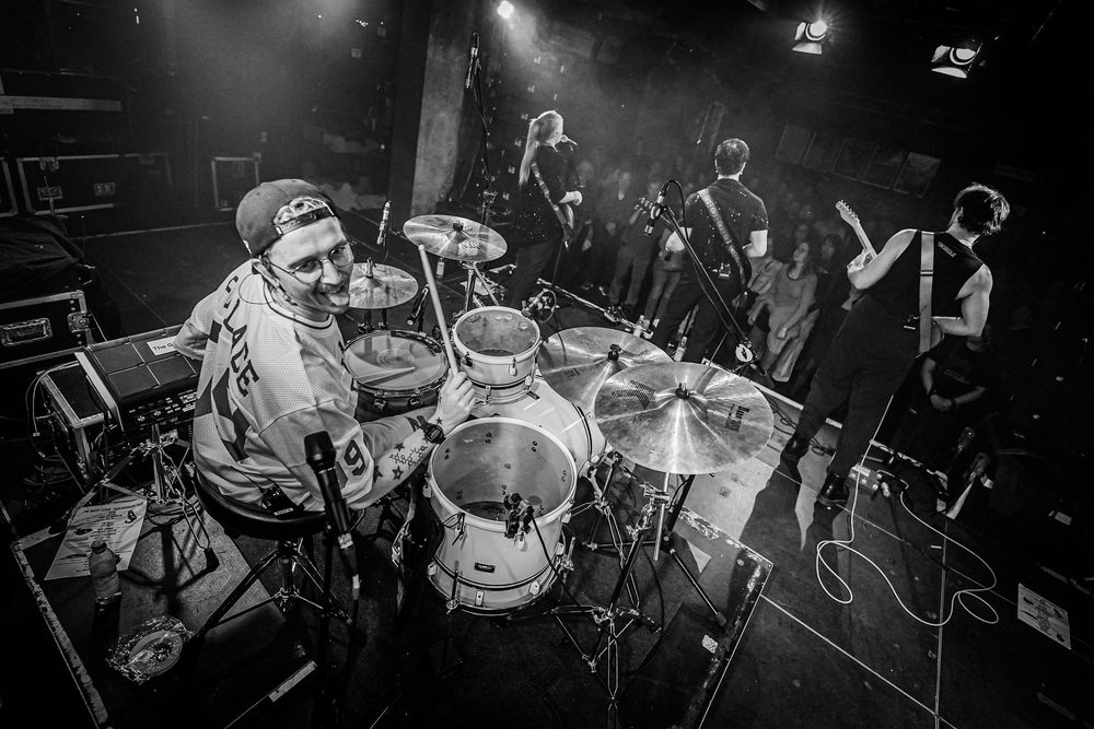 Black and white photo of a drummer performing on stage with band members in background at a live music venue.