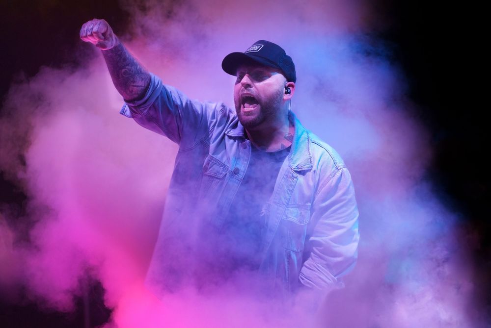 Male performer raises fist on smoky stage illuminated by pink and blue lights during energetic concert.