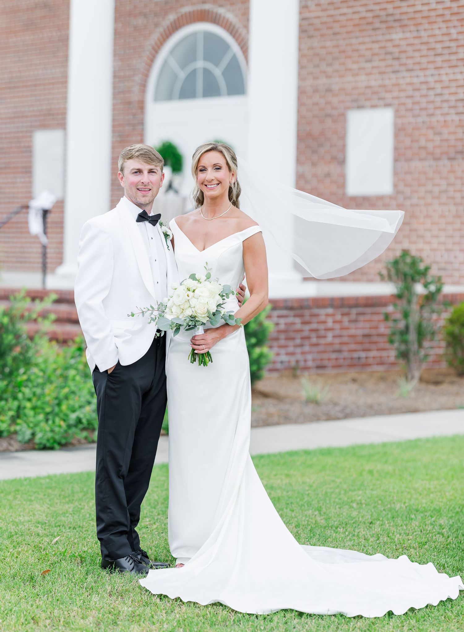 Bride and groom pose outside brick church, she holds white bouquet in elegant gown, he wears white tuxedo jacket.