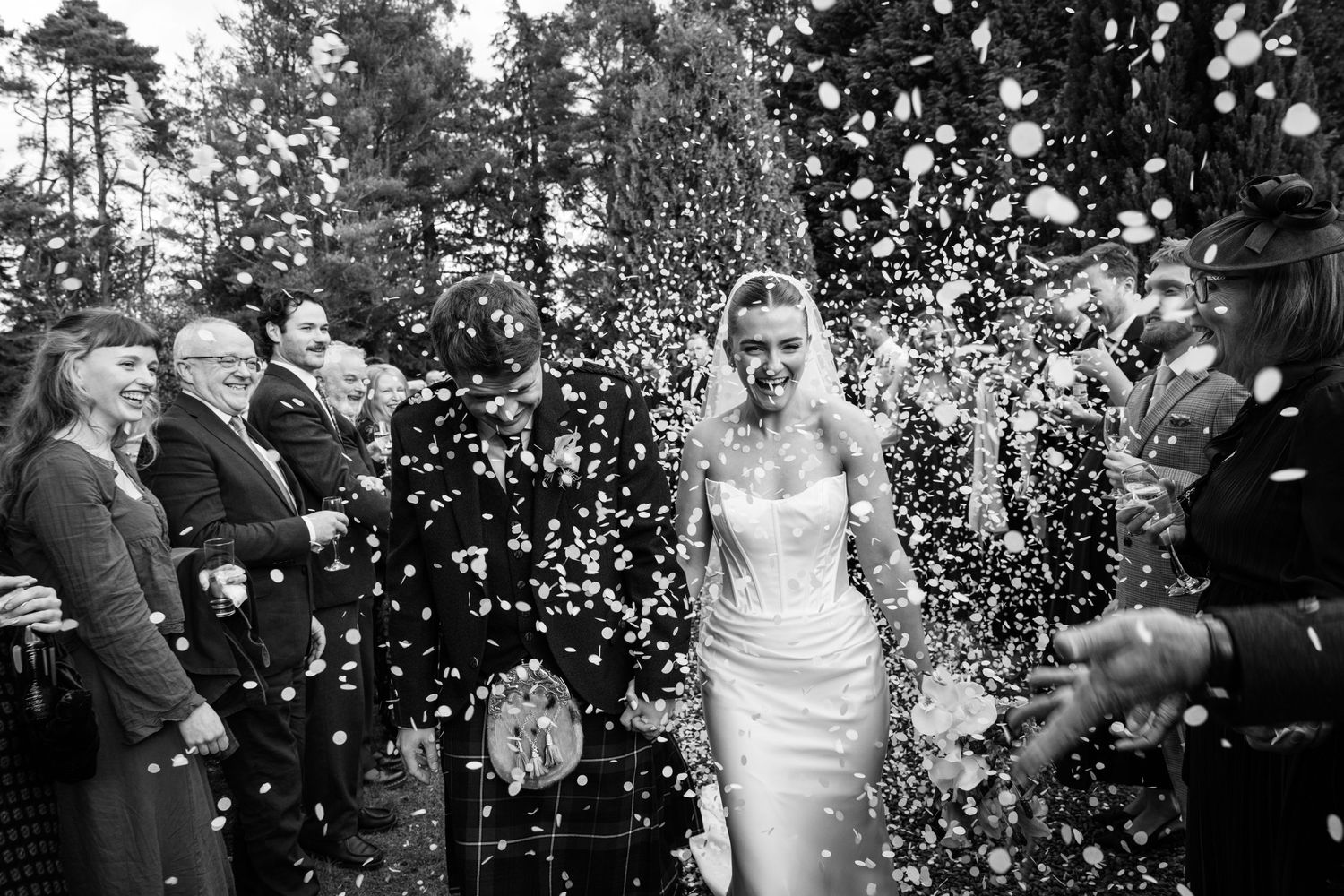 Bride walks through confetti shower in elegant white gown, surrounded by smiling guests, black and white wedding photography.