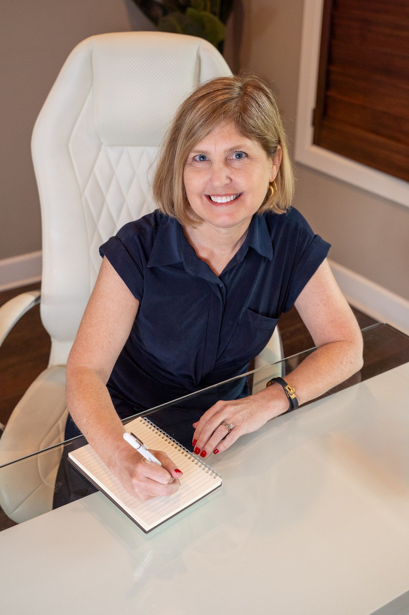 Smiling professional woman in navy dress writing in notebook at glass desk in modern office.