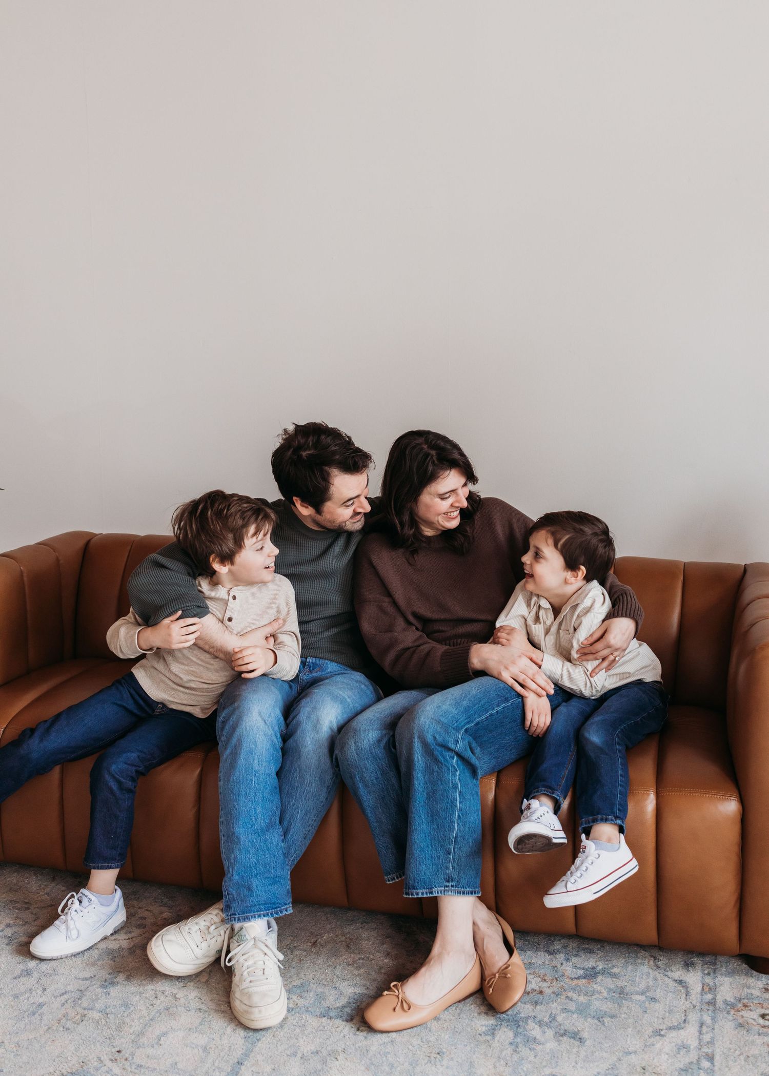 A happy family of four sits together on a brown leather couch, smiling and laughing in a cozy living room.