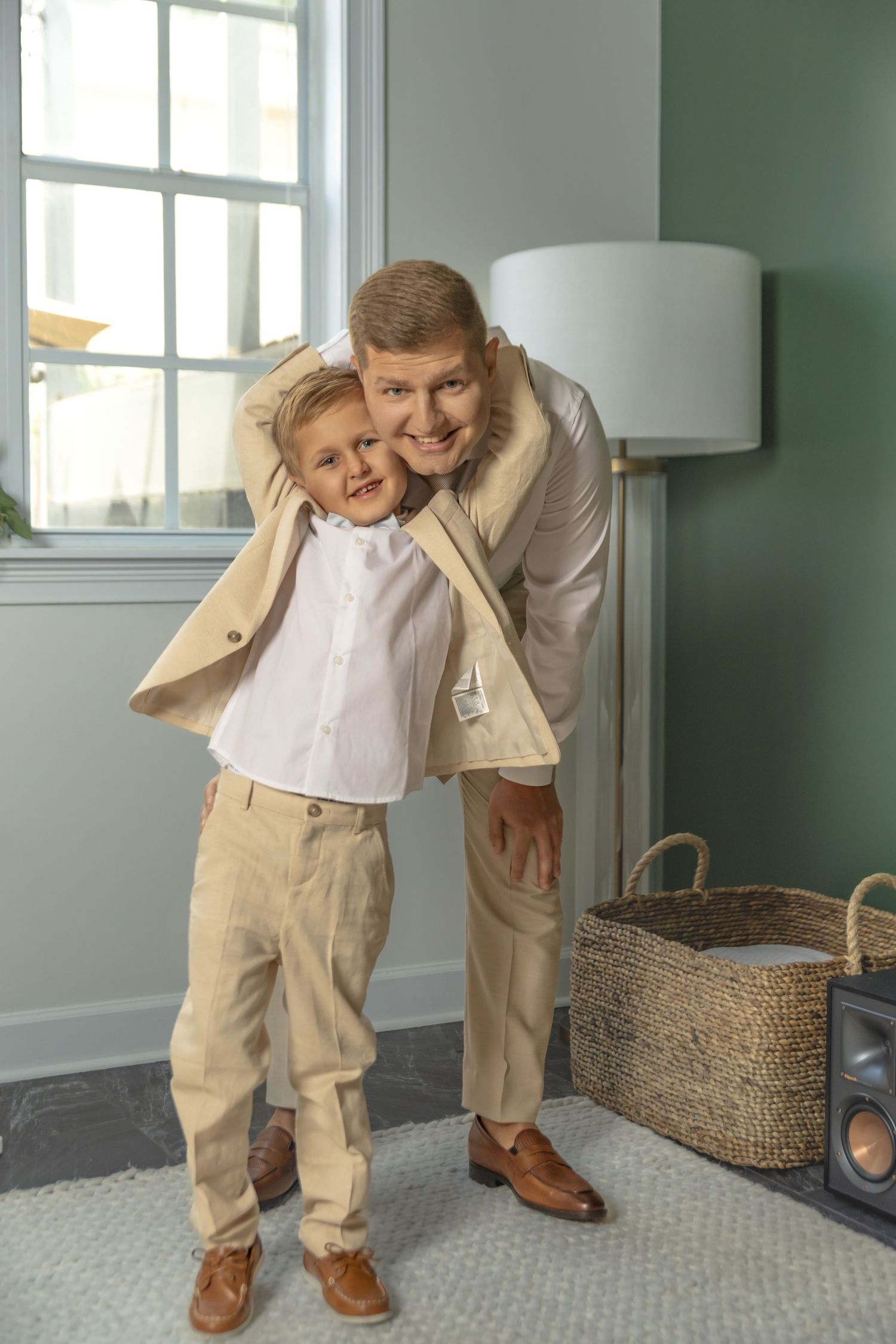A man and young boy in matching beige outfits share a warm hug indoors near a window.