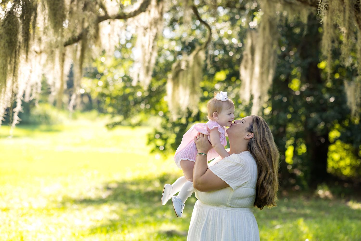 A mother in a white dress lifts her baby girl under moss-draped oak trees in a sunlit park.