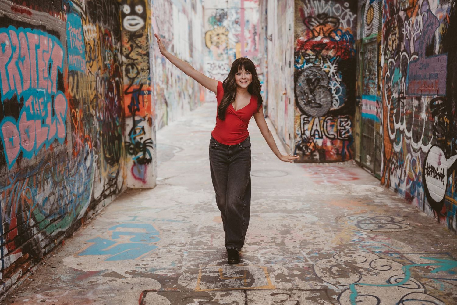 Woman in red top and black jeans posing in a colorful graffiti-covered alleyway with arms outstretched.