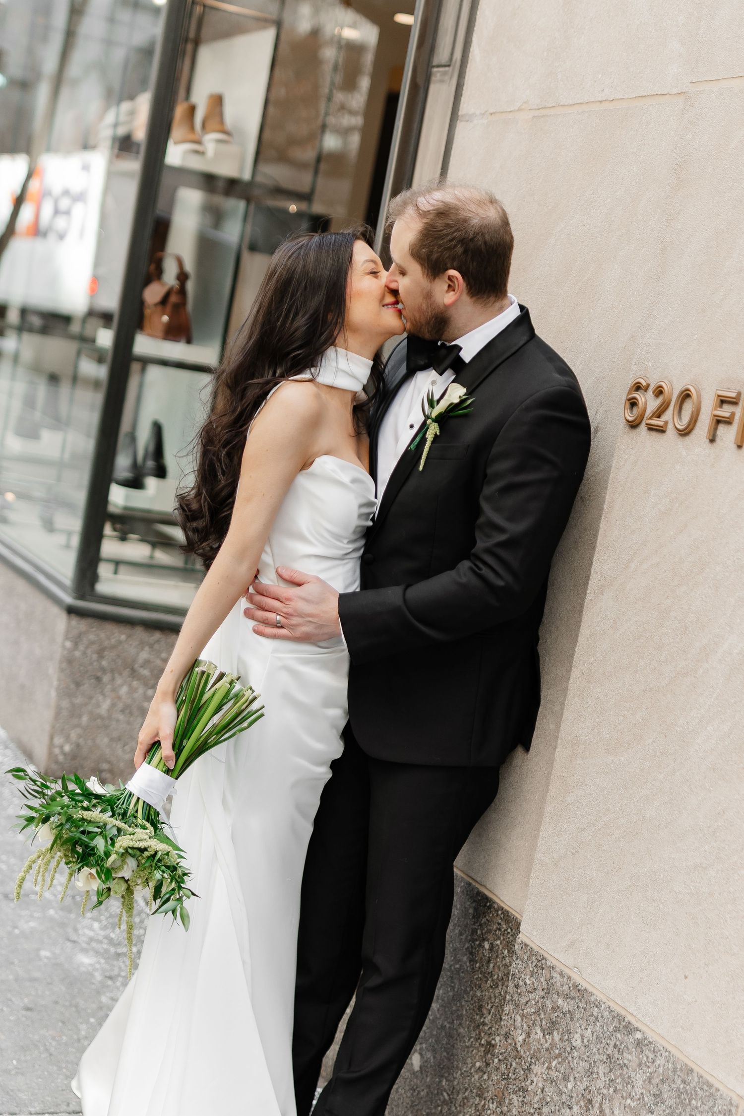 Bride and groom share a romantic kiss outdoors against a stone building, bride holding a green and white bouquet.