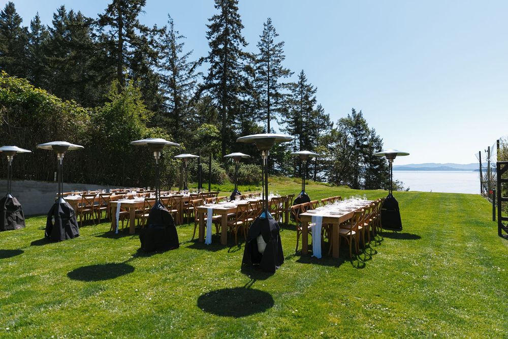 Elegant outdoor wedding reception setup with long tables, umbrellas, and heaters on a lush green lawn overlooking the water.