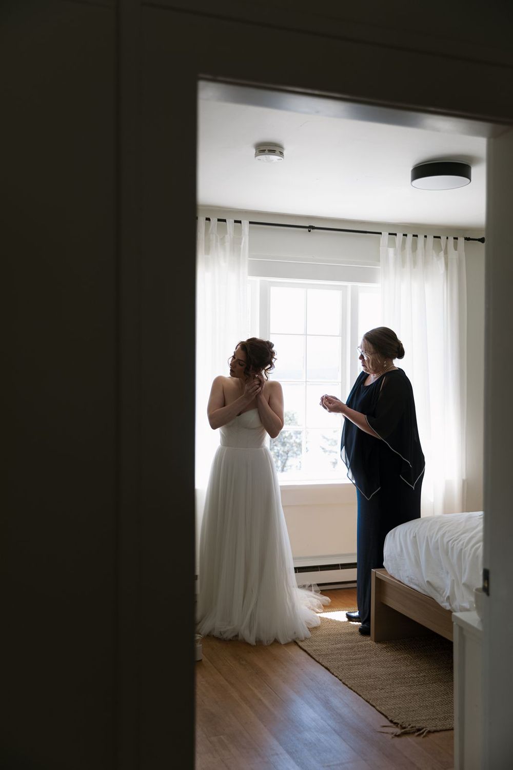 Bride in white gown stands with guest by sunlit window in bedroom during wedding preparations.